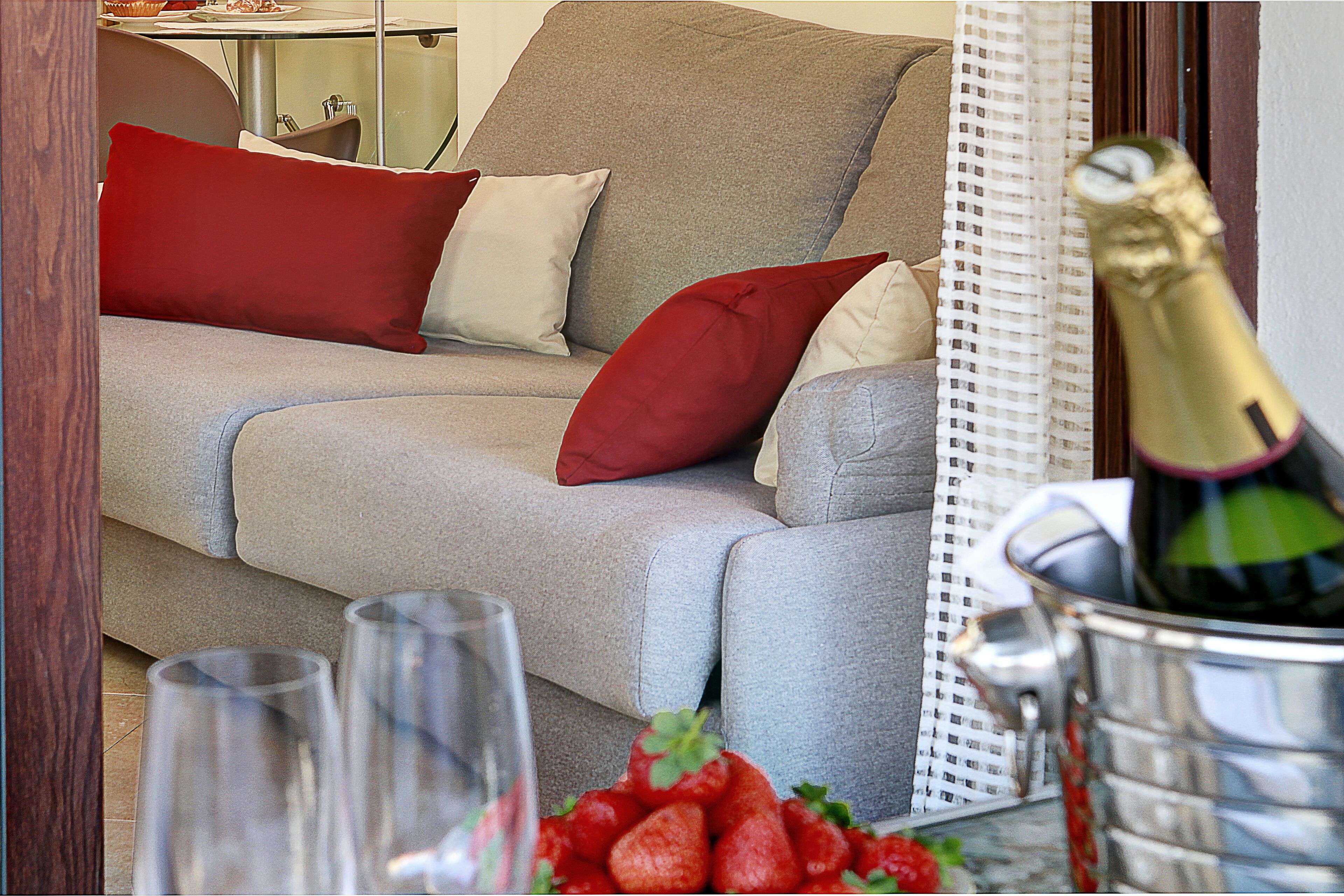 Un canapé gris avec des coussins rouges et blancs, une table en verre sur laquelle se trouvent une bouteille de champagne et des verres, et un panier de fraises.