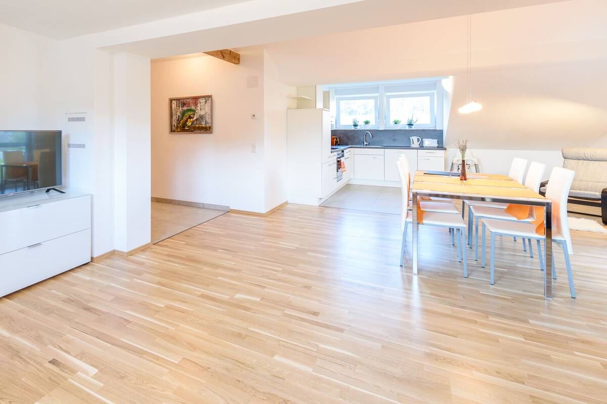 A modern living room with a flat-screen TV, a dining area with a table and chairs, and a kitchen with a window.
