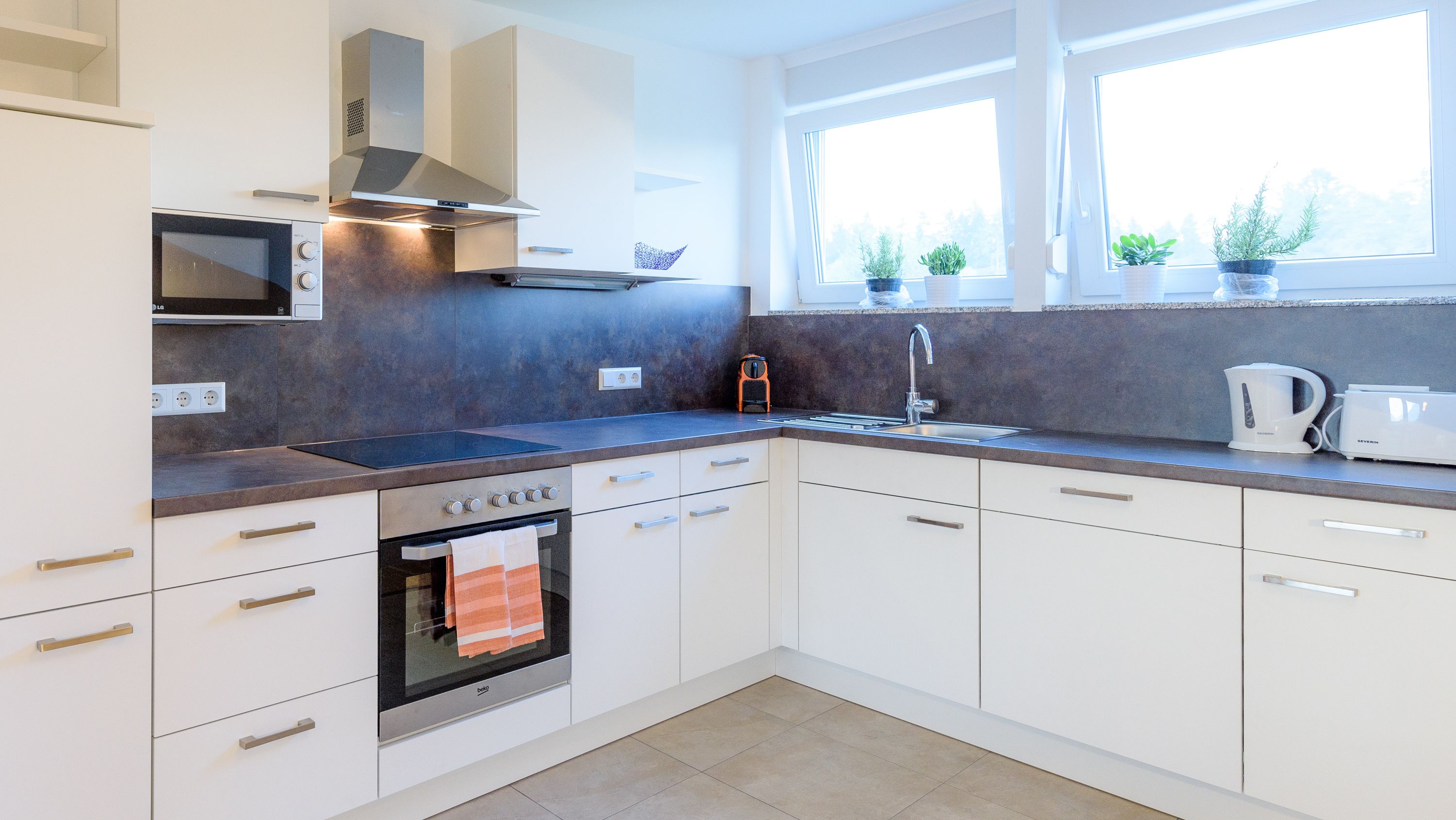A modern kitchen with white cabinets, a built-in oven, a microwave, a sink, and a window with potted plants.