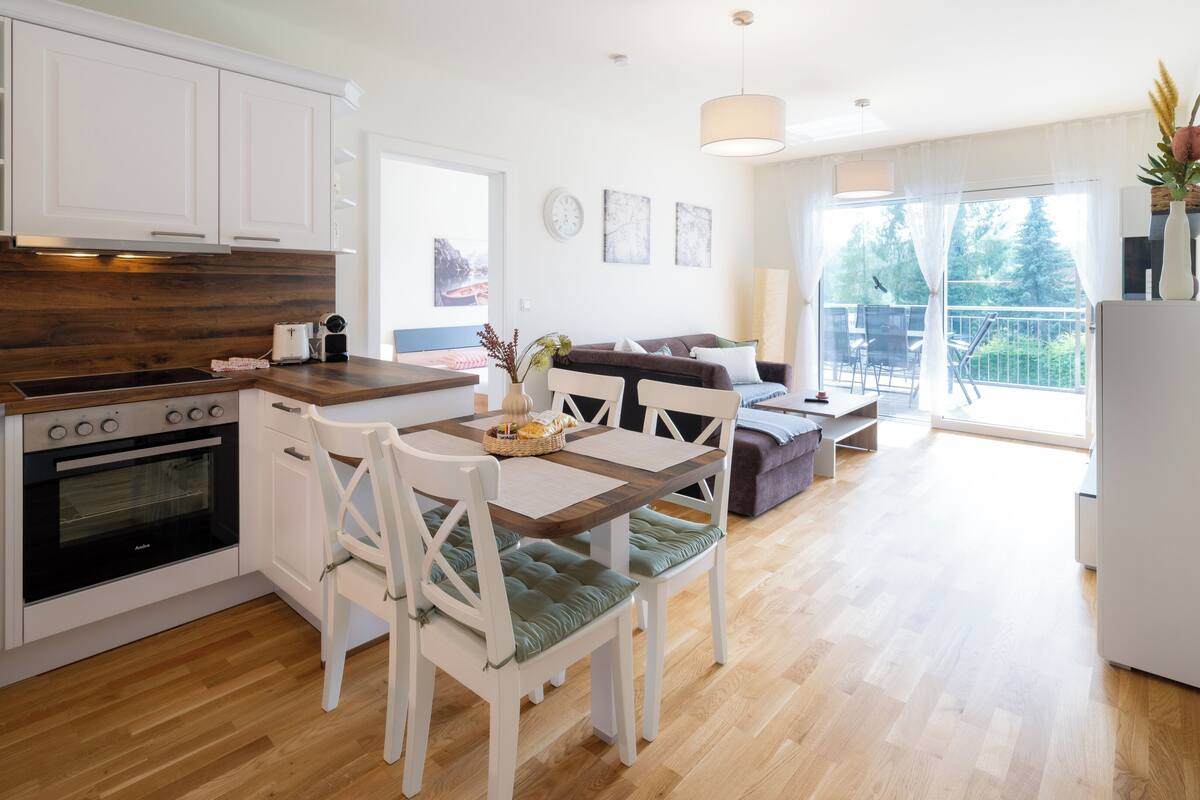 A modern kitchen with white cabinets, a wooden countertop, and a built-in oven. There is a dining area with a wooden table and white chairs.