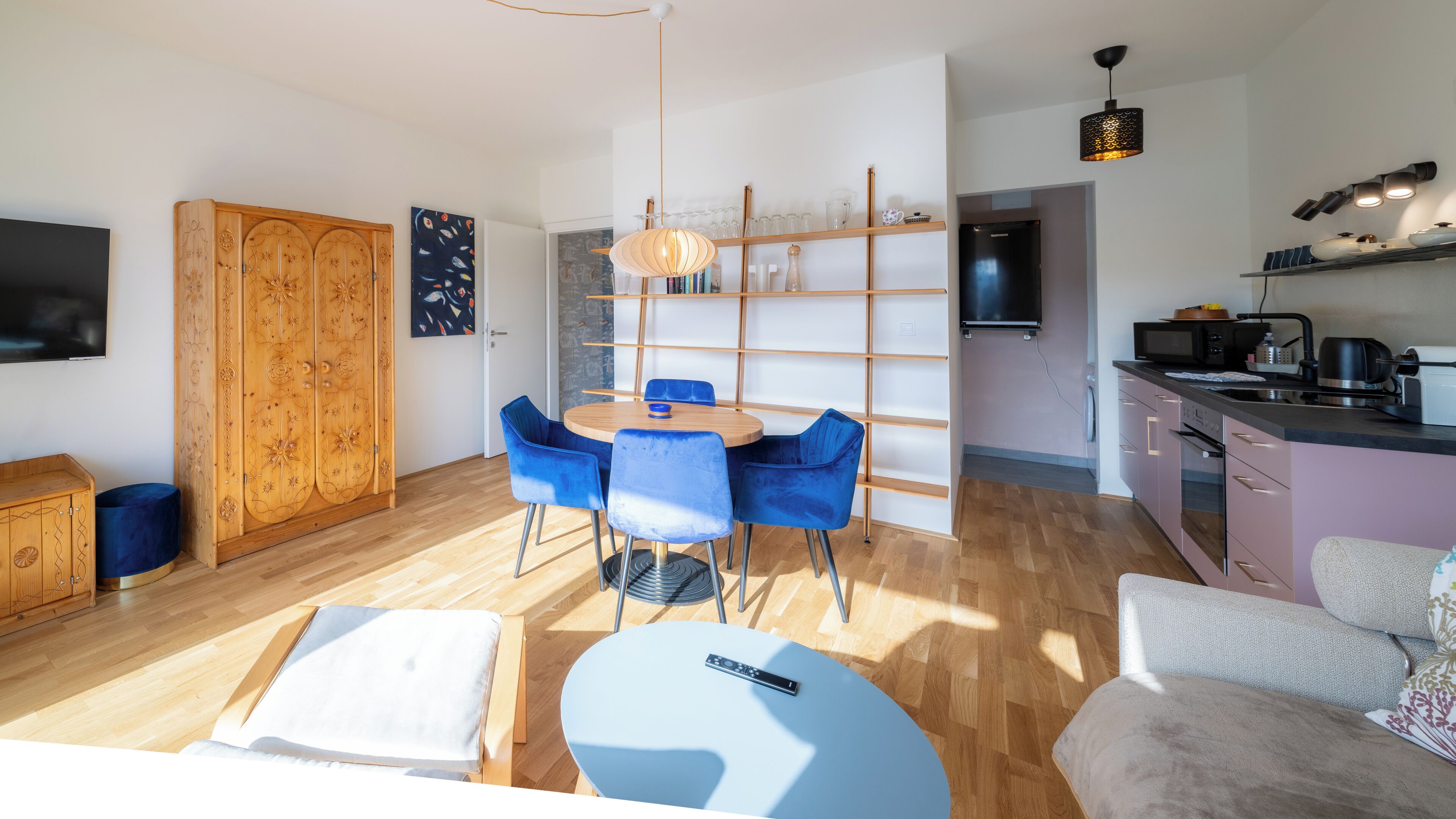 A modern living room with a wooden wardrobe, a dining table with blue chairs, a TV, and a kitchen area with a microwave and kettle.