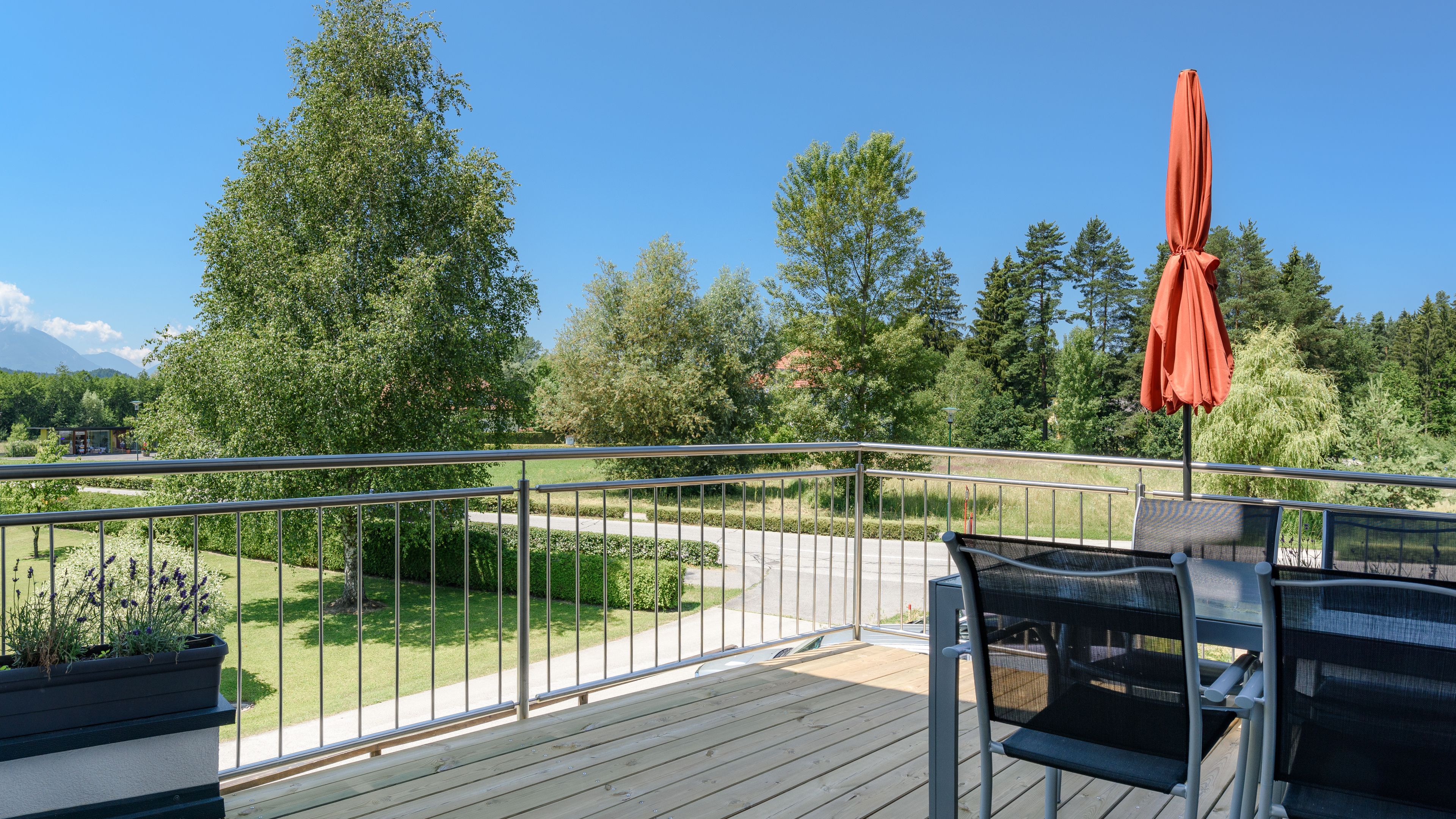Eine Holzterrasse mit Metallgeländer, Sitzgelegenheiten im Freien und Blick auf einen angelegten Garten mit Bäumen und einem Weg.