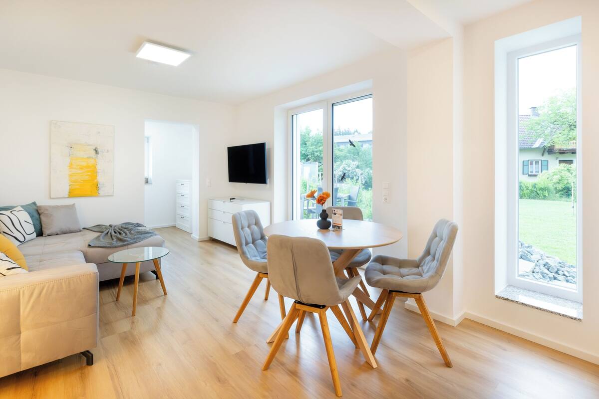 A modern living room with a sofa, a dining table with chairs, a flat-screen TV mounted on the wall, and large windows offering a view of the outdoors.