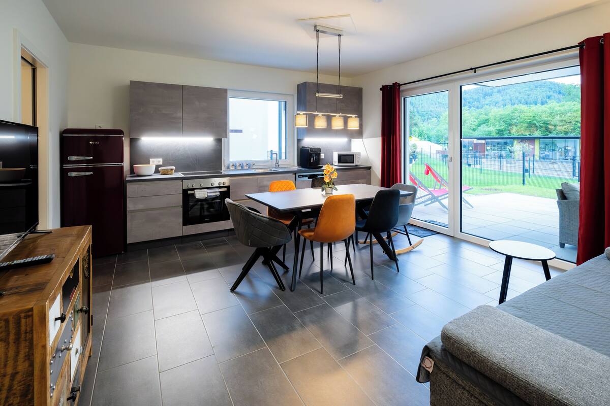 A modern kitchen with a dining area, a sofa, and a view of a green landscape.