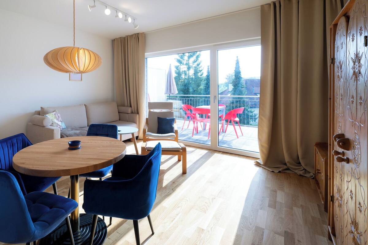 A modern living room with a round wooden table, blue chairs, a sofa, and a balcony with red chairs.