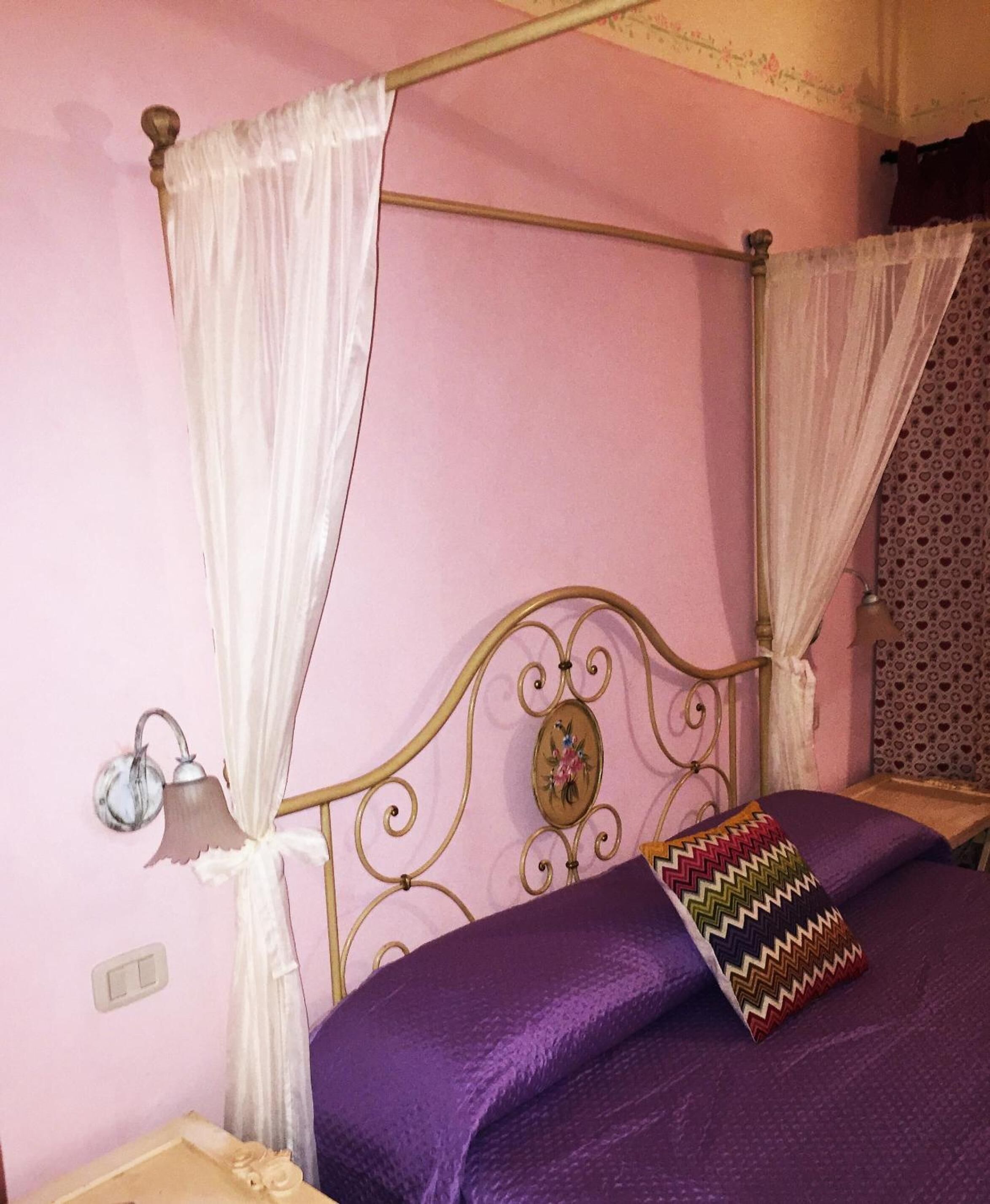Un lit avec une tête de lit en métal, des rideaux blancs et une couvre-lit violette.