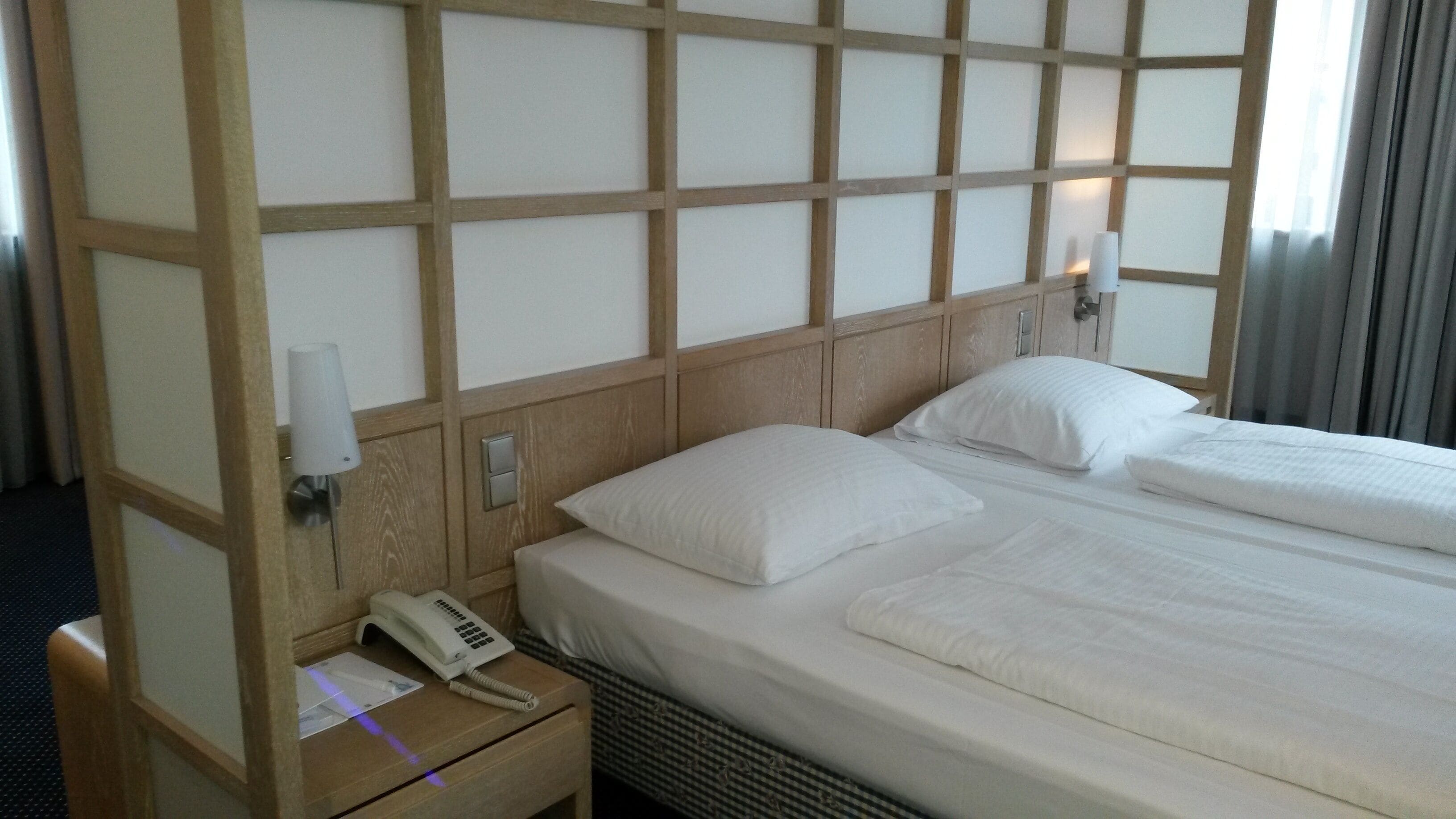 Habitación de hotel con cabecera de madera, teléfono en la mesita de noche y cama con ropa blanca bien tendida.