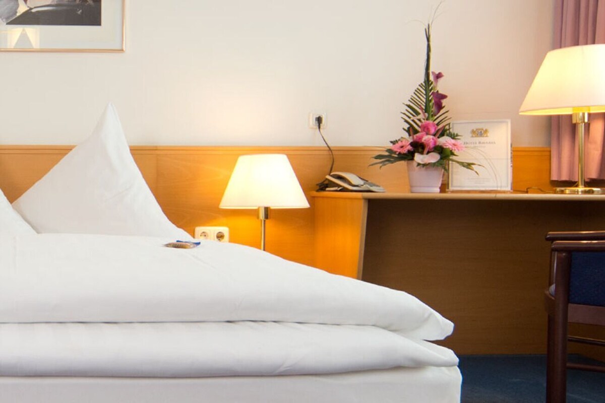 A hotel room with a bed, two bedside lamps, a desk lamp on a desk, a chair, and a framed picture on the wall.