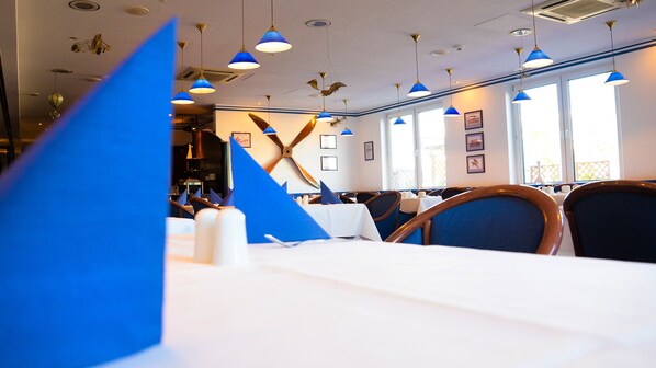 Un interior de restaurante con lámparas colgantes azules, manteles blancos y cuadros en las paredes.