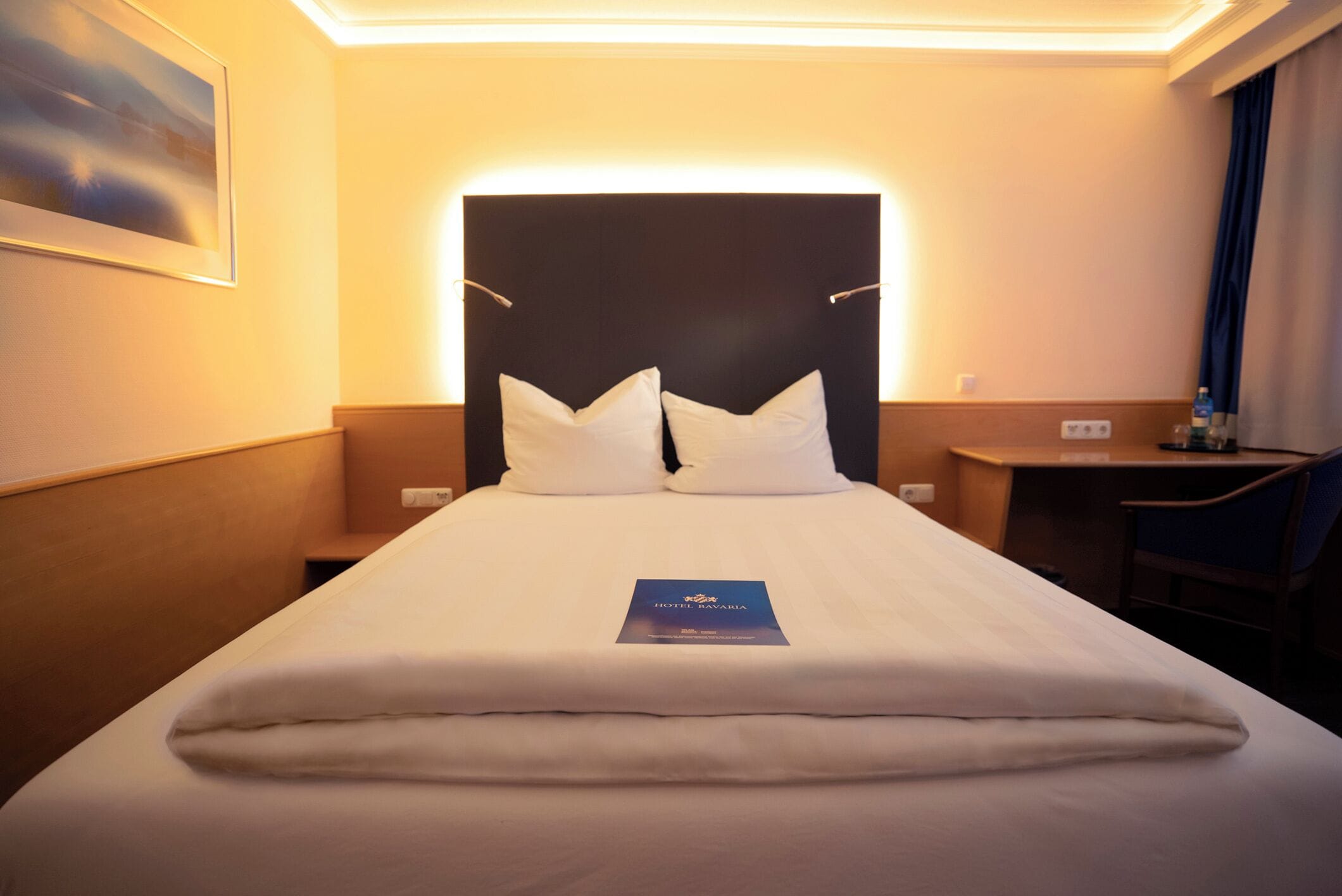 A hotel room with a neatly made bed, a headboard with integrated lighting, a small bedside table, and a desk with a chair.