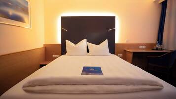 A hotel room with a neatly made bed, a headboard with integrated lighting, a small bedside table, and a desk with a chair.