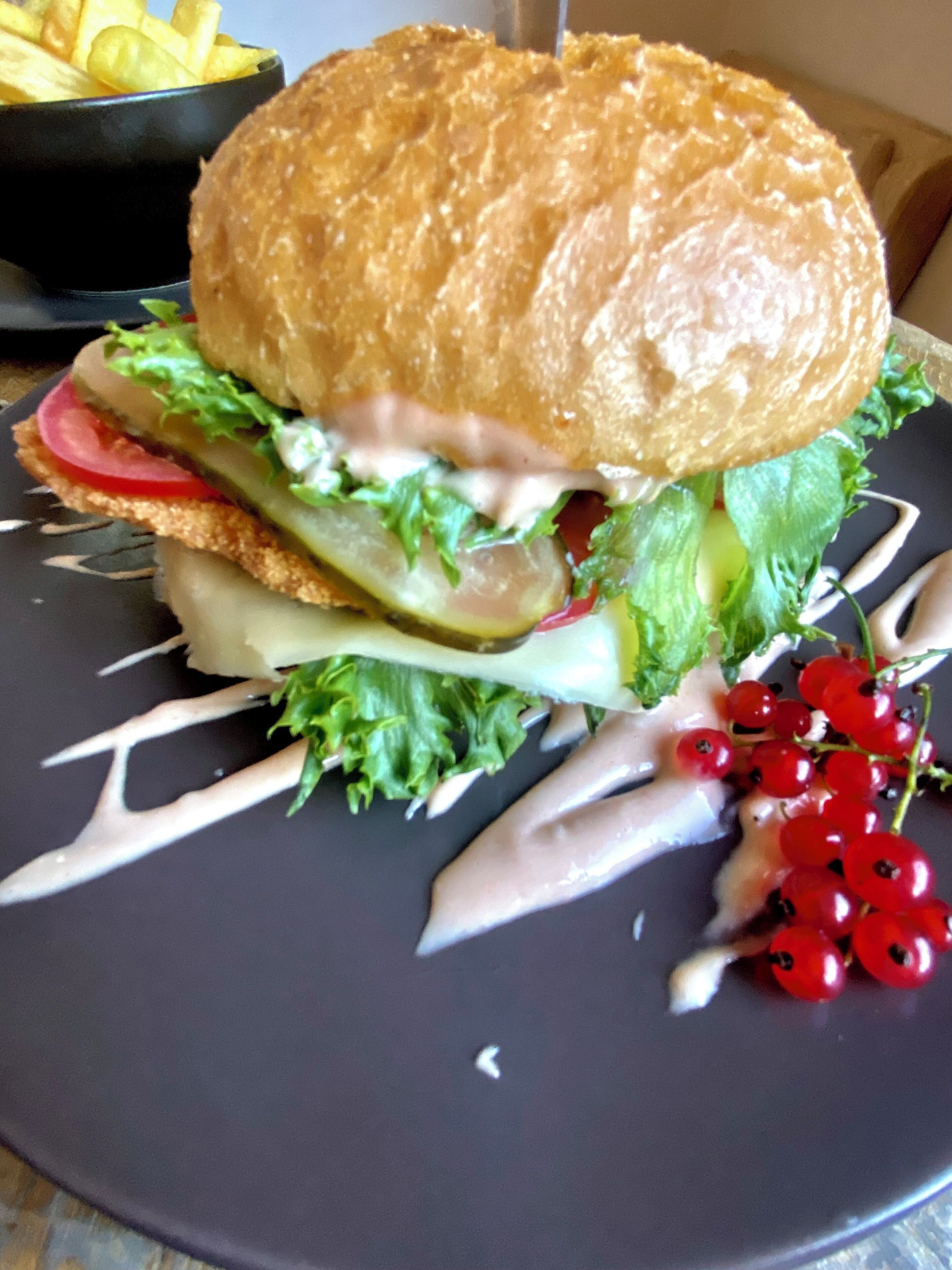 Um hambúrguer com alface, tomate e molho cremoso, servido em um prato escuro, acompanhado de frutas vermelhas e legumes em conserva.