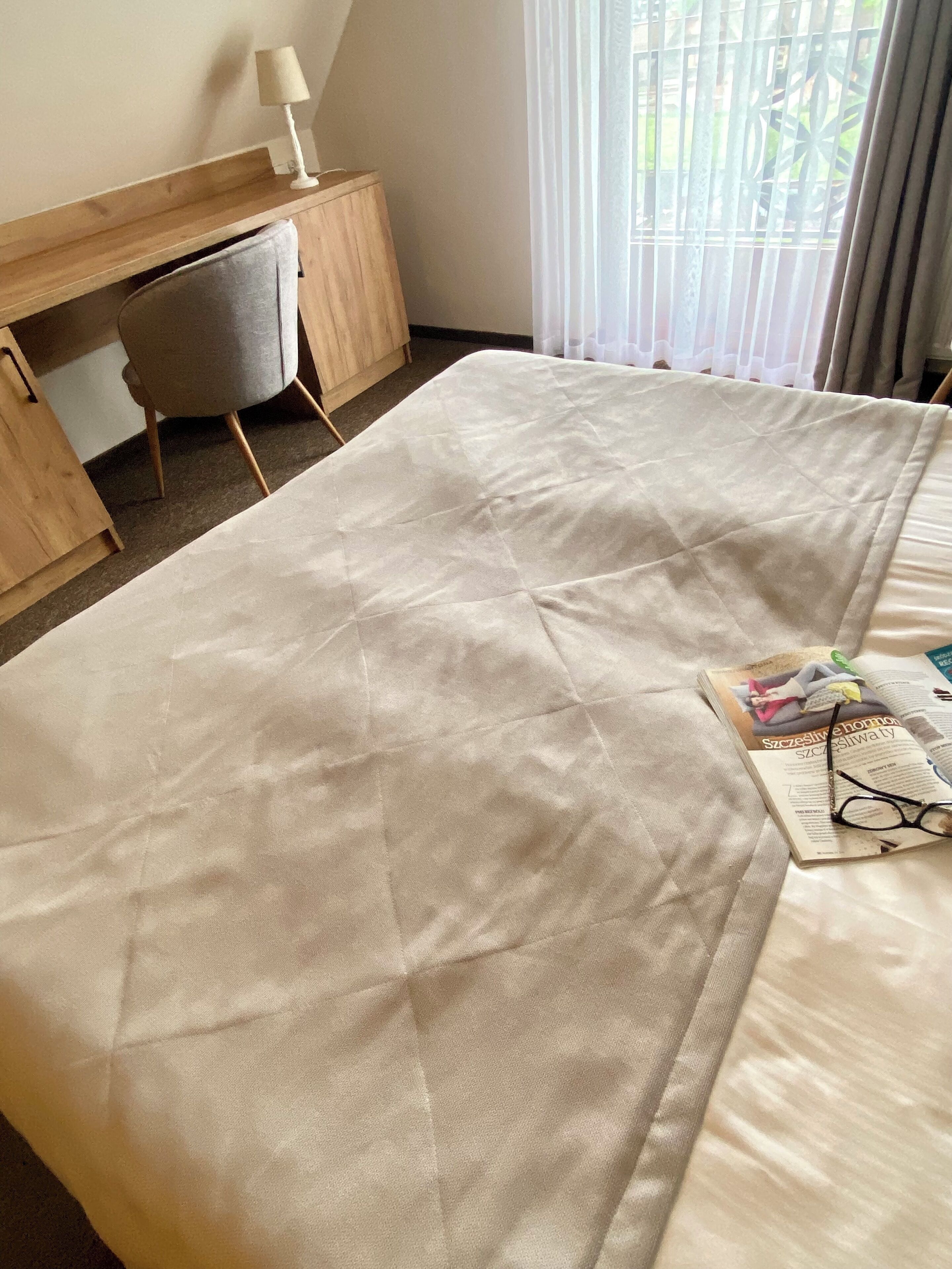 A neatly made bed with a quilted cover, a small desk with a lamp, and a chair.