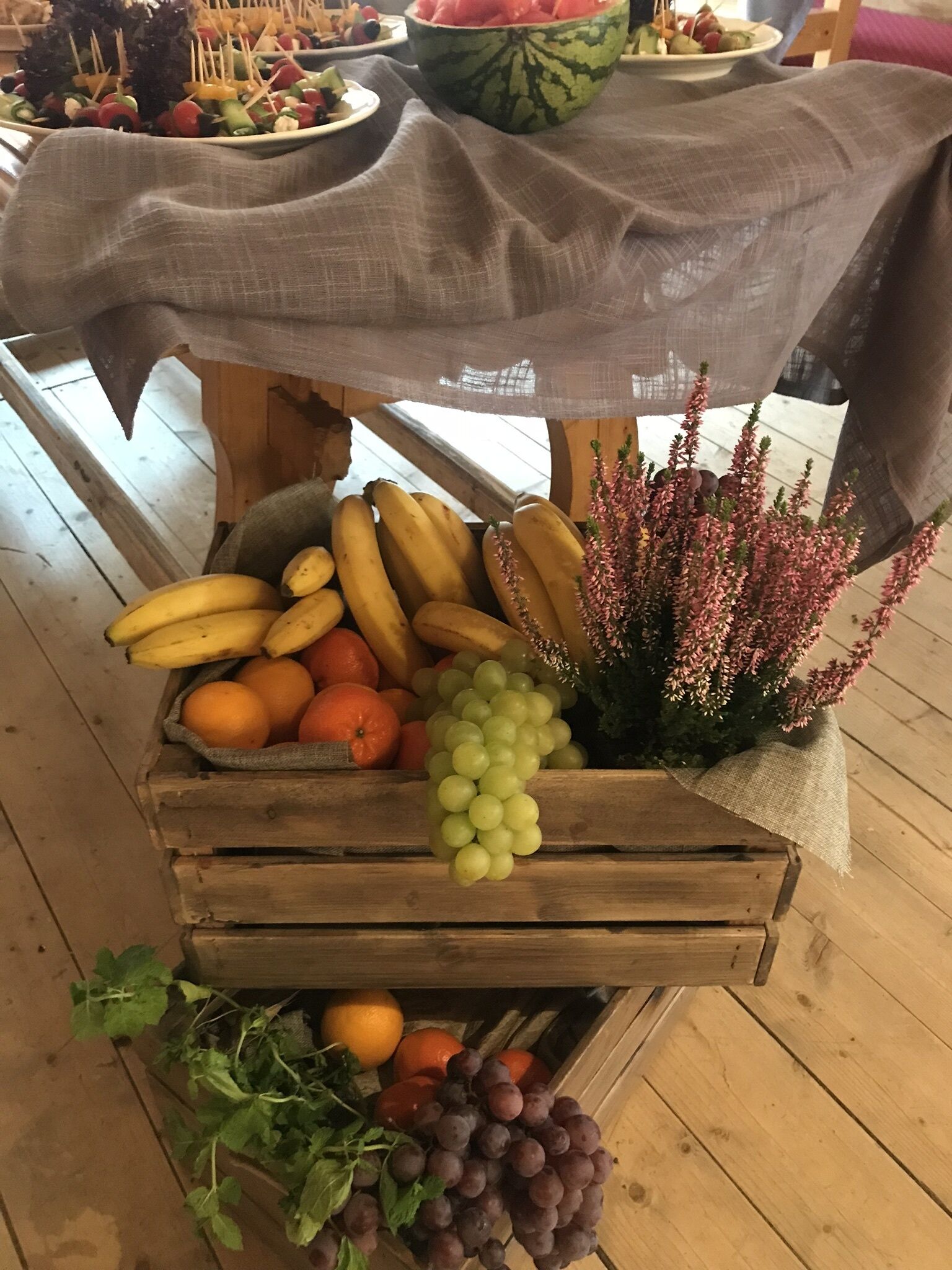 Una exhibición de madera con frutas y verduras, incluyendo plátanos, naranjas, uvas y hierbas aromáticas.