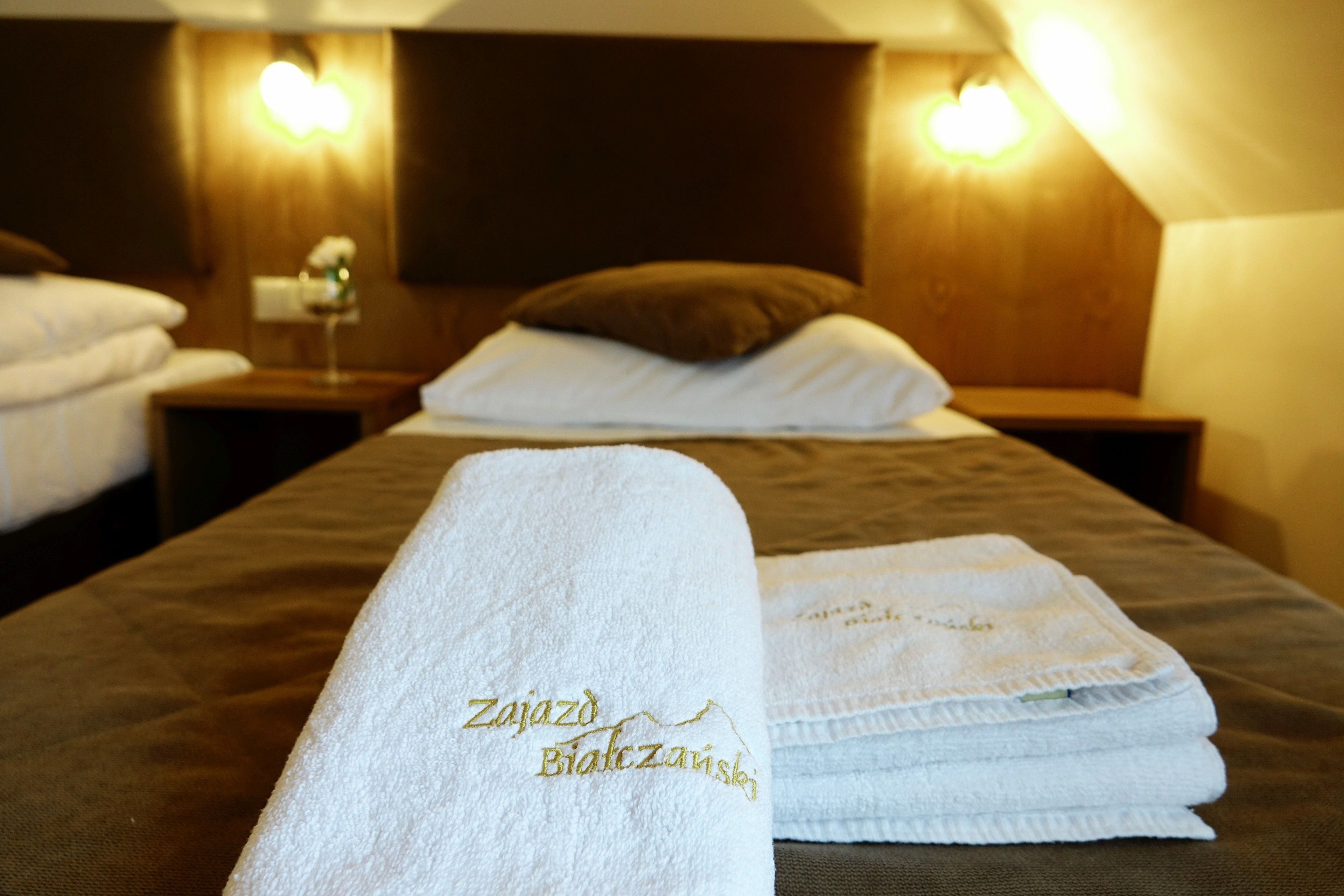 Habitación de hotel con toallas ordenadamente dobladas sobre la cama, cabecero de madera y lámparas fijadas a la pared.