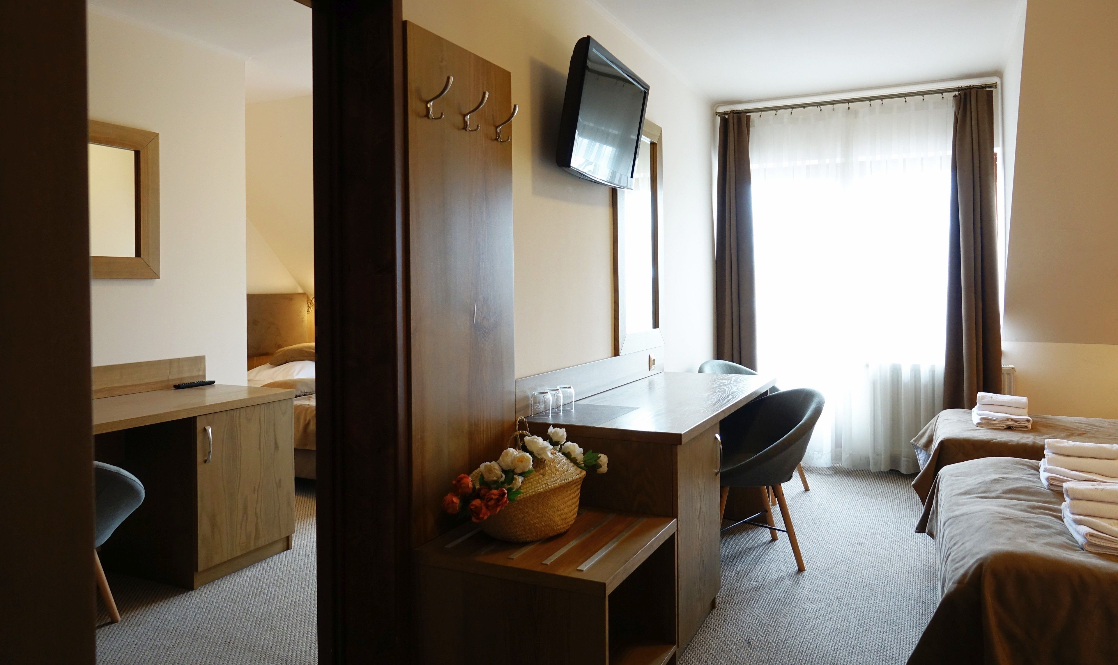 A hotel room with a wooden desk, a television mounted on the wall, a bed with a brown bedspread, and a window with curtains.