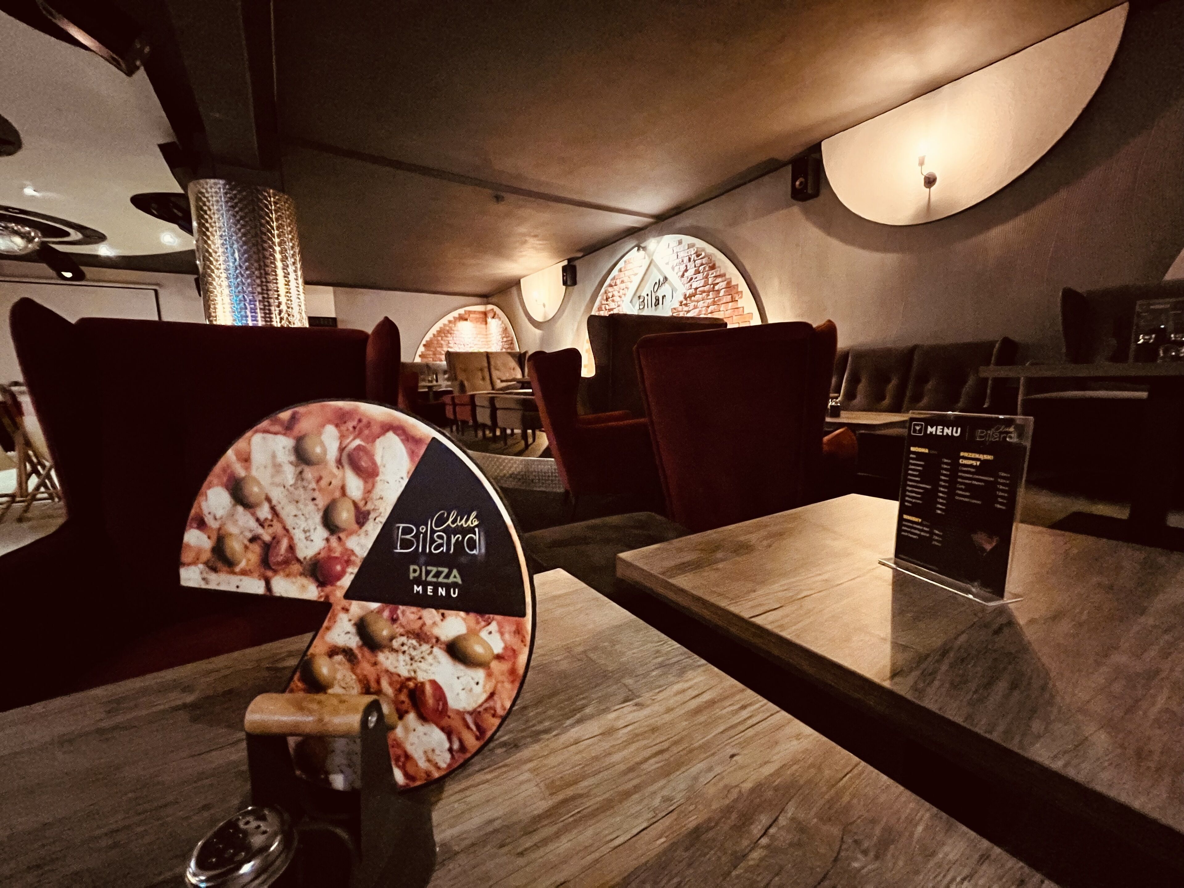 Een Club Bialard pizza menu in een restaurant, met houten tafels en stoelen, een bakstenen muur en een menustandaard.