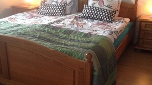Una cama de madera con colcha verde y almohadas estampadas, una mesita de noche con lámpara y un aparador de madera.