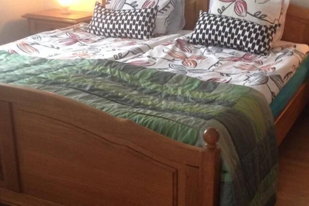 Un lit en bois avec une couette verte et des oreillers à motifs, une table de chevet avec une lampe et une commode en bois.