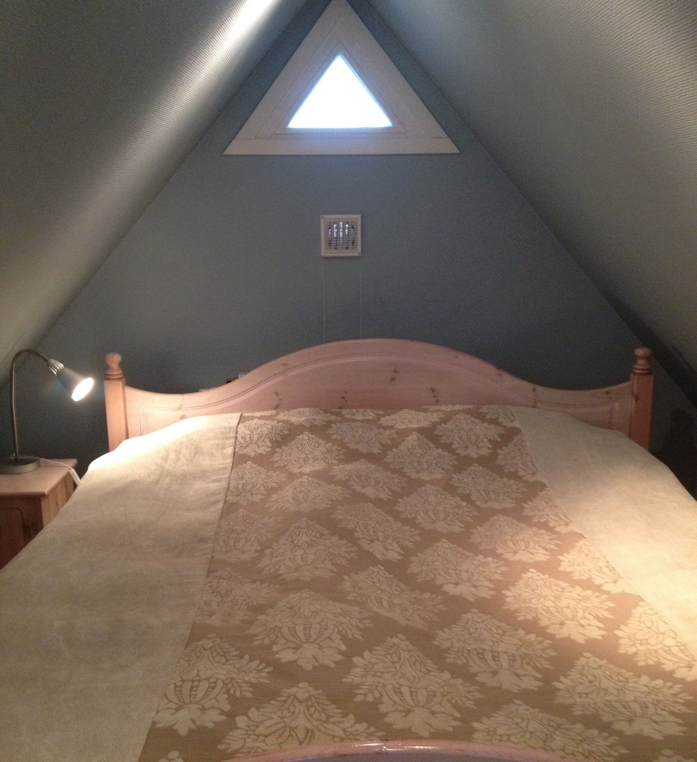 Un dormitorio con techo inclinado, un tragaluz triangular, una cama con colcha estampada, una lámpara de noche y una pequeña rejilla de ventilación en la pared.