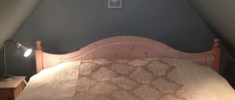 Un dormitorio con techo inclinado, un tragaluz triangular, una cama con colcha estampada, una lámpara de noche y una pequeña rejilla de ventilación en la pared.