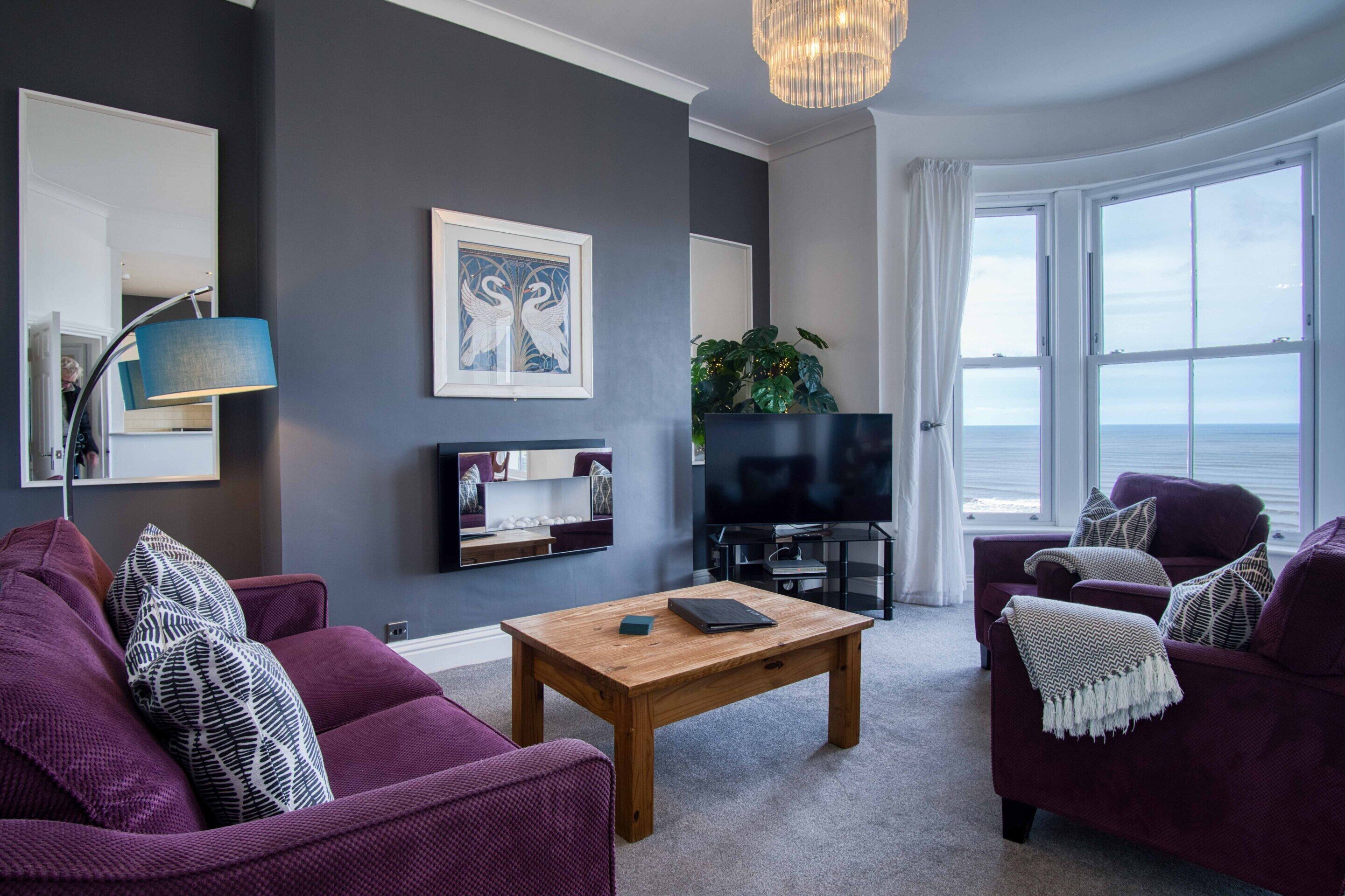 Ein modernes Wohnzimmer mit dunkler Wand, einem Holztisch, violetten Sofas, einem Fernseher und einem großen Fenster mit Aussicht.