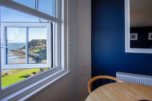 Une pièce dotée d’une grande fenêtre donnant sur un paysage côtier, d’une table en bois et d’un mur bleu orné de tableaux encadrés.