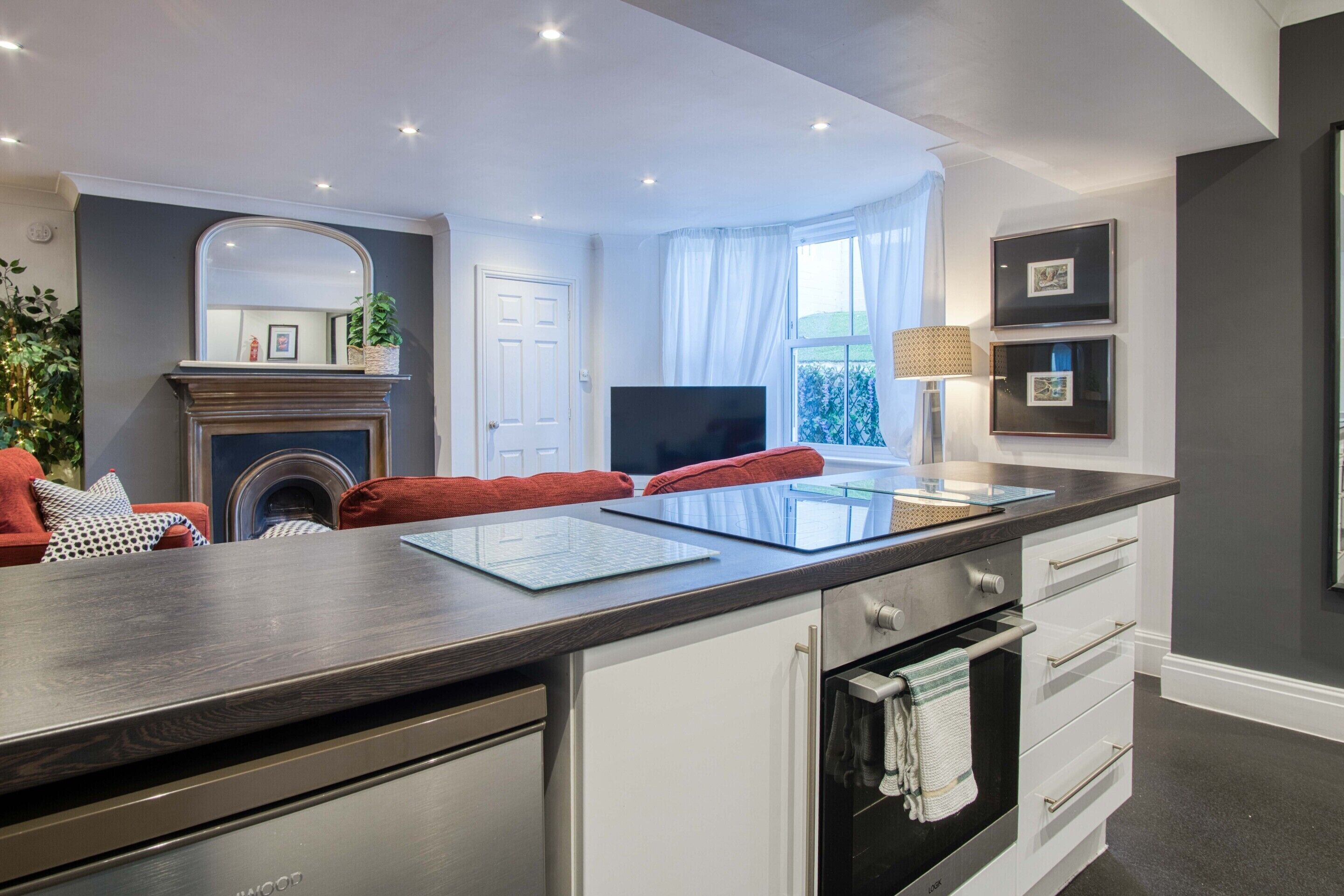 Een moderne keuken met een donker aanrecht, witte kasten en een ingebouwde oven. Er is een open haard met een spiegel erboven, een televisie en een raam met gordijnen.
