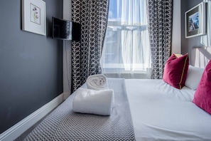 Une chambre d’hôtel avec un lit, un téléviseur fixé au mur et une fenêtre ornée de rideaux à motifs.