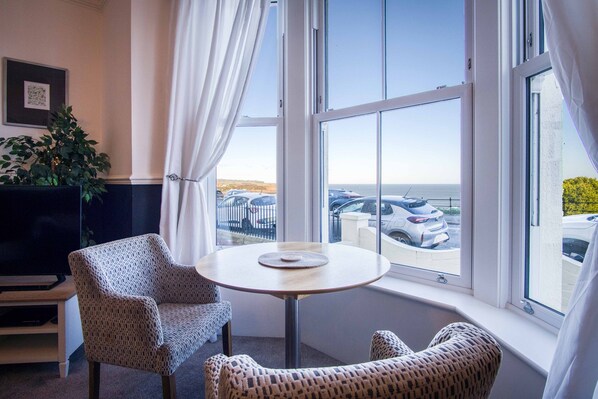 A room with a round table, two patterned chairs, a TV, and a large window with a view of parked cars and a body of water. - Blue Horizon Apartments at The Helaina (Scarborough)