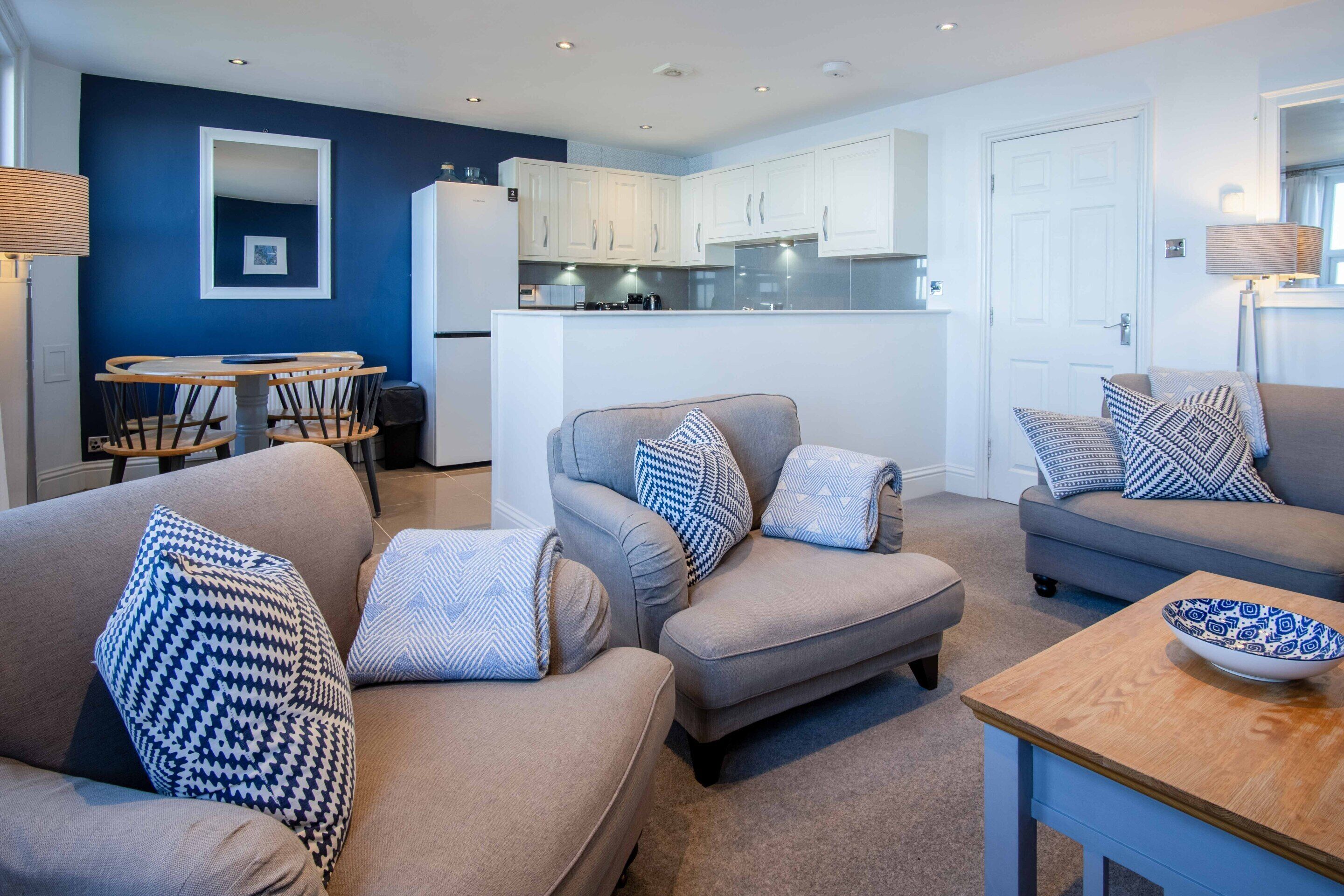 A modern living room with a grey sofa set, a wooden coffee table, and a kitchen area in the background.