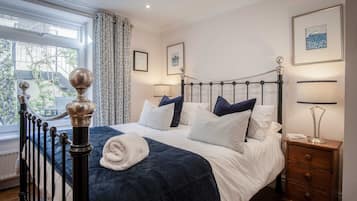 A neatly made bed with a dark blue comforter and white linens, a bedside table with a lamp, and a window with patterned curtains.