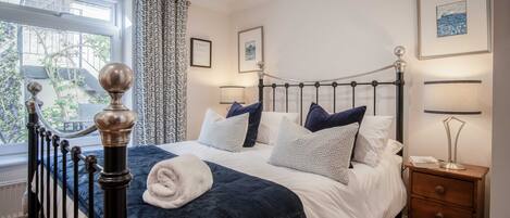A neatly made bed with a dark blue comforter and white linens, a bedside table with a lamp, and a window with patterned curtains.
