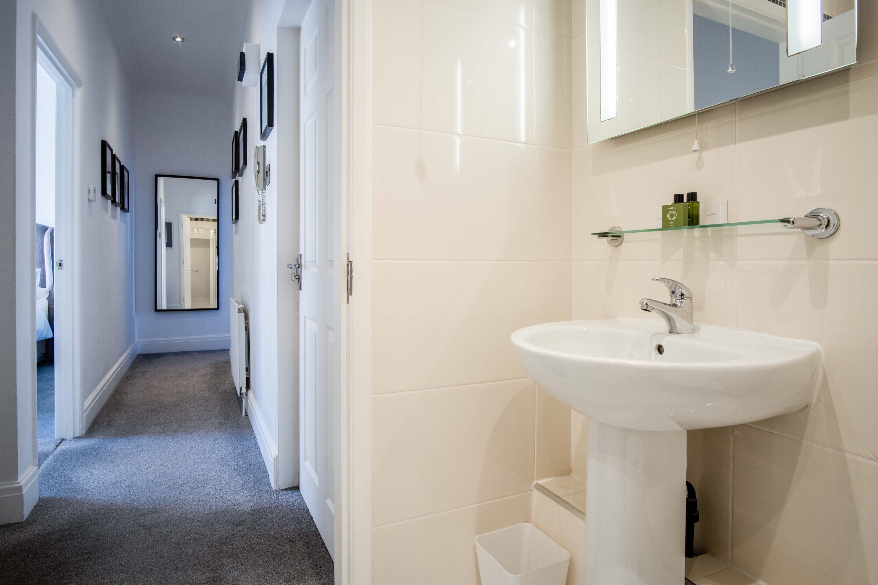 A hallway leading to a bathroom with a white sink, a mirror, and a glass shelf with toiletries.