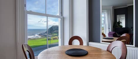 A dining area with a wooden table and chairs, large windows with a view of the sea and hills, and a modern interior design.