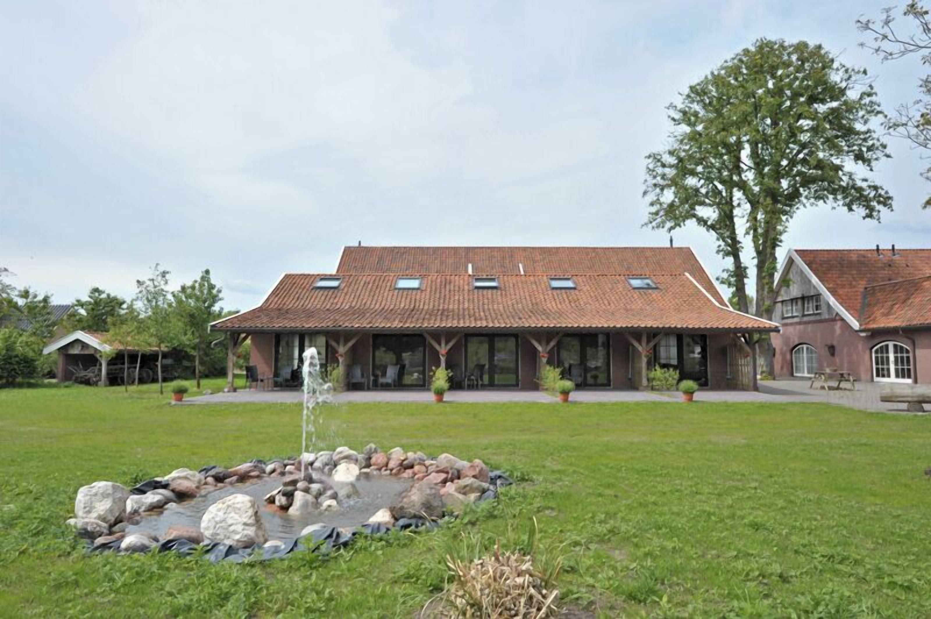 Een groot huis met een pannendak, een terras en een kleine vijver met rotsen en een fontein.