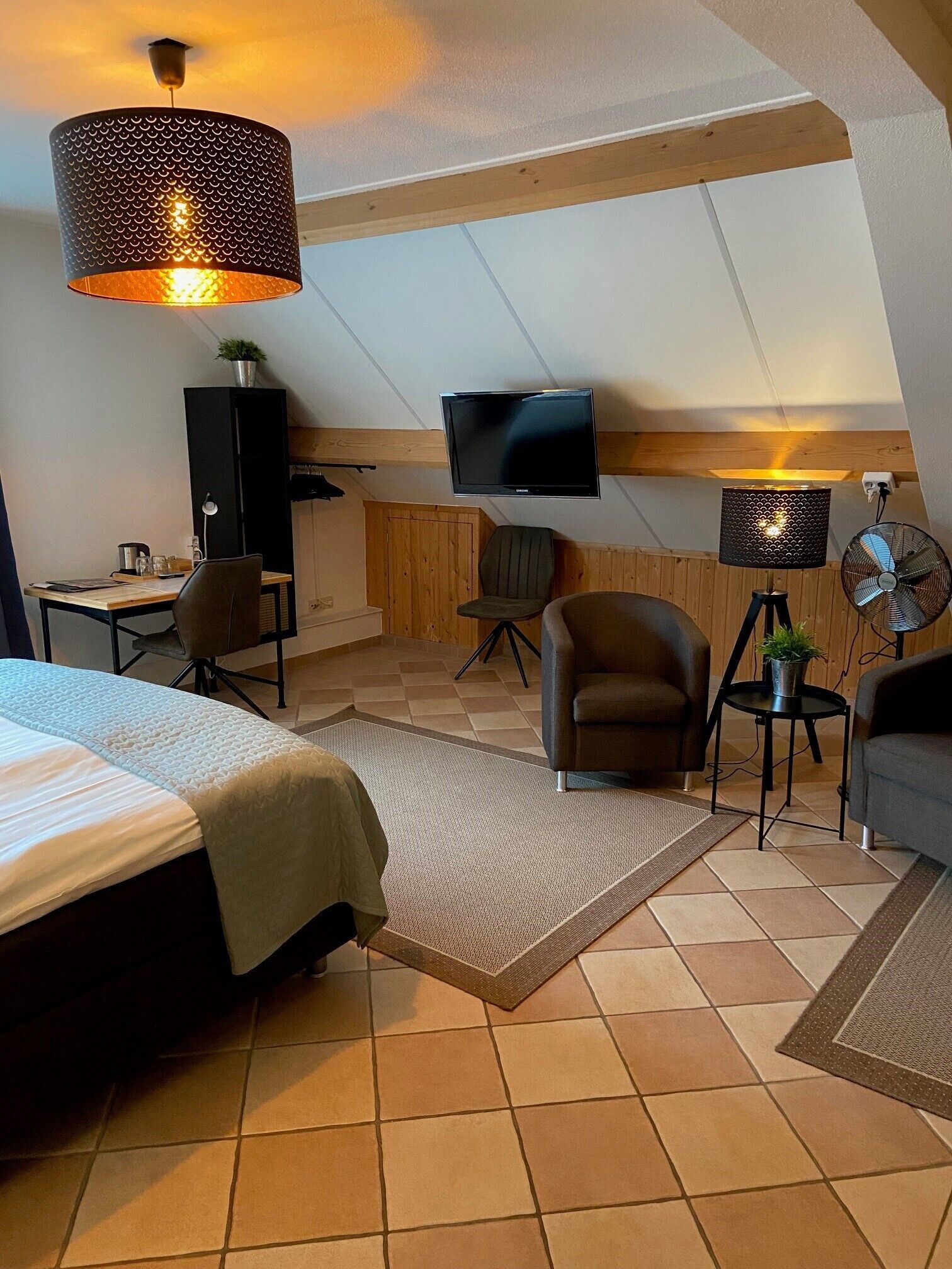 A cozy, well-lit attic bedroom with a bed, a desk, a chair, a TV, and a fan.