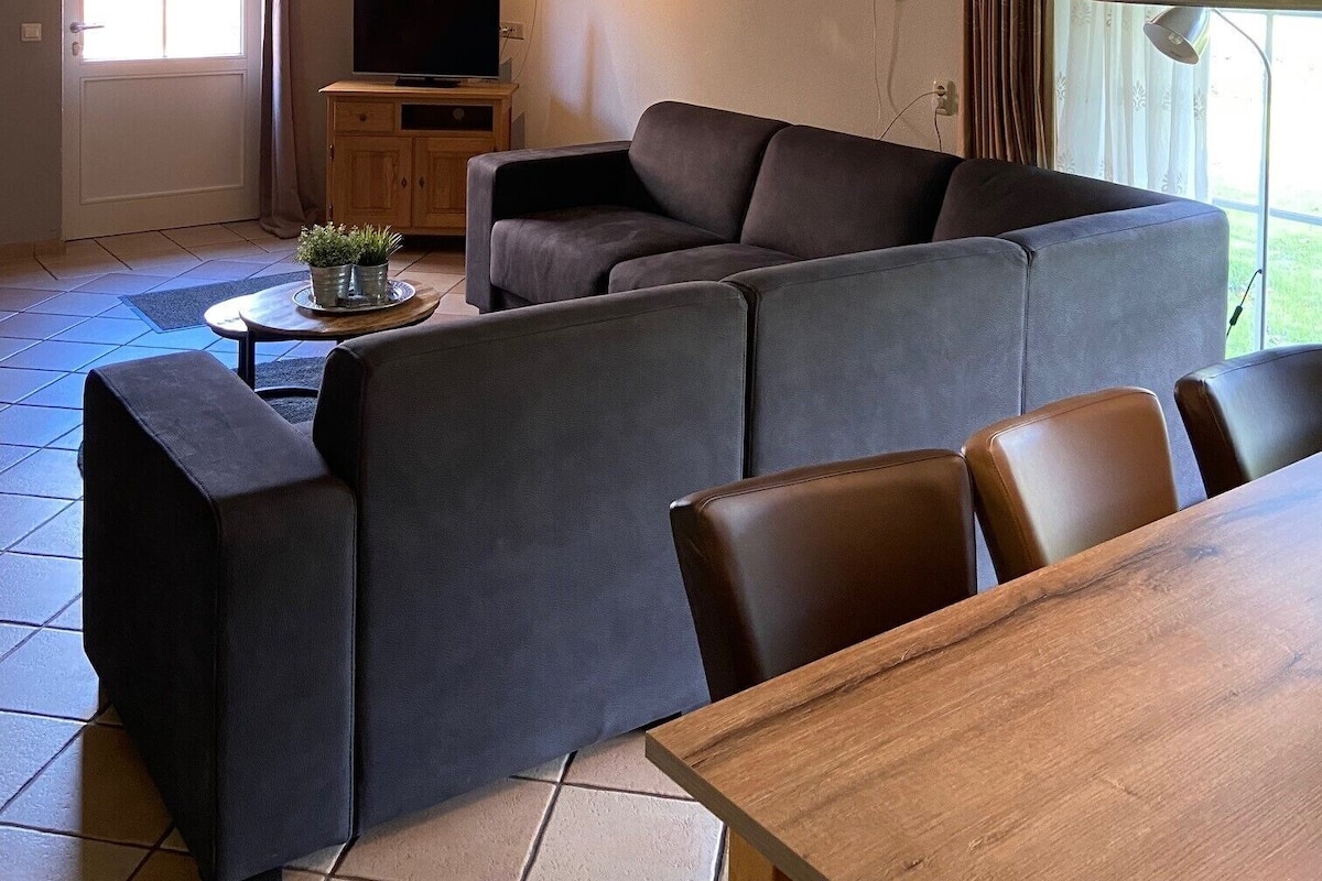 A living room with a sectional sofa, a wooden dining table, a television, and a window with curtains.