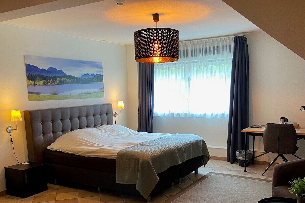 A bedroom with a large bed, a desk, a chair, a lamp, and a view of a lake through the window.