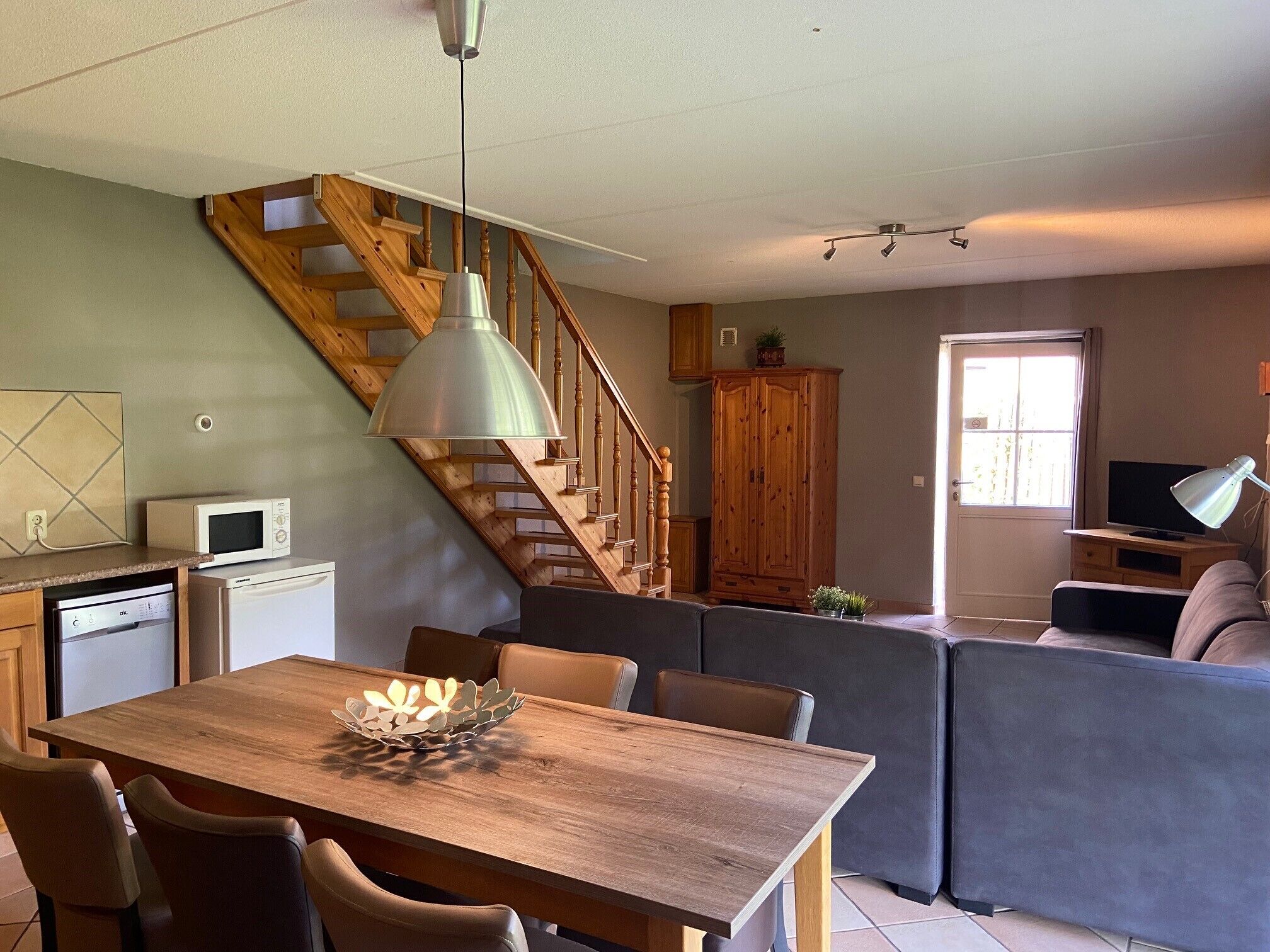 Una cocina moderna con una escalera de madera, un comedor con mesa y sillas de madera, y una sala de estar con sofá y televisor.