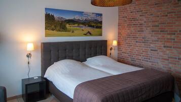 A bedroom with a large bed, two bedside lamps, a painting of a mountain landscape, and a brick wall.