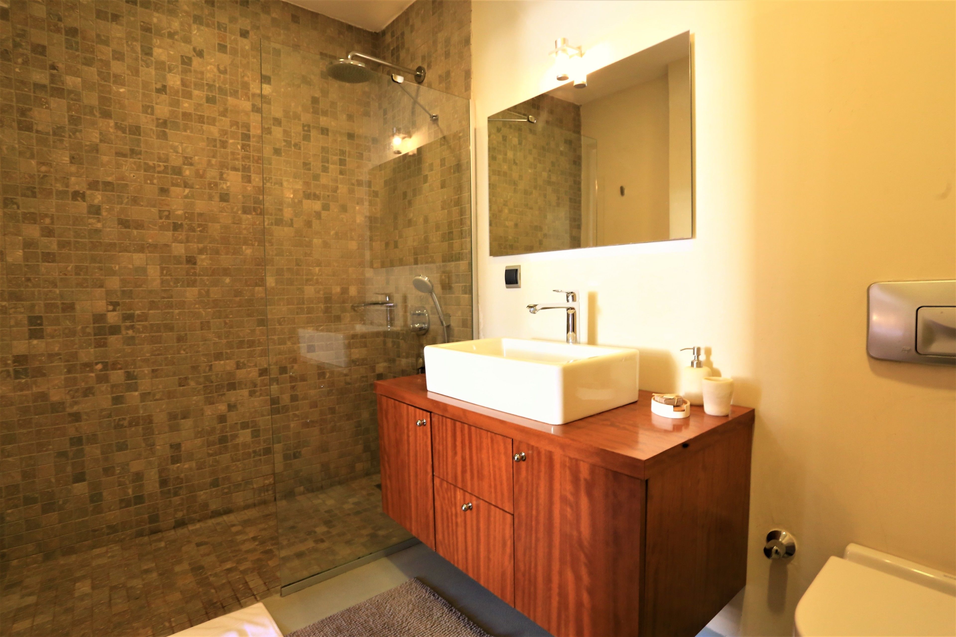 Un baño con pared de ducha azulejada, tocador de madera con lavamanos y un inodoro.