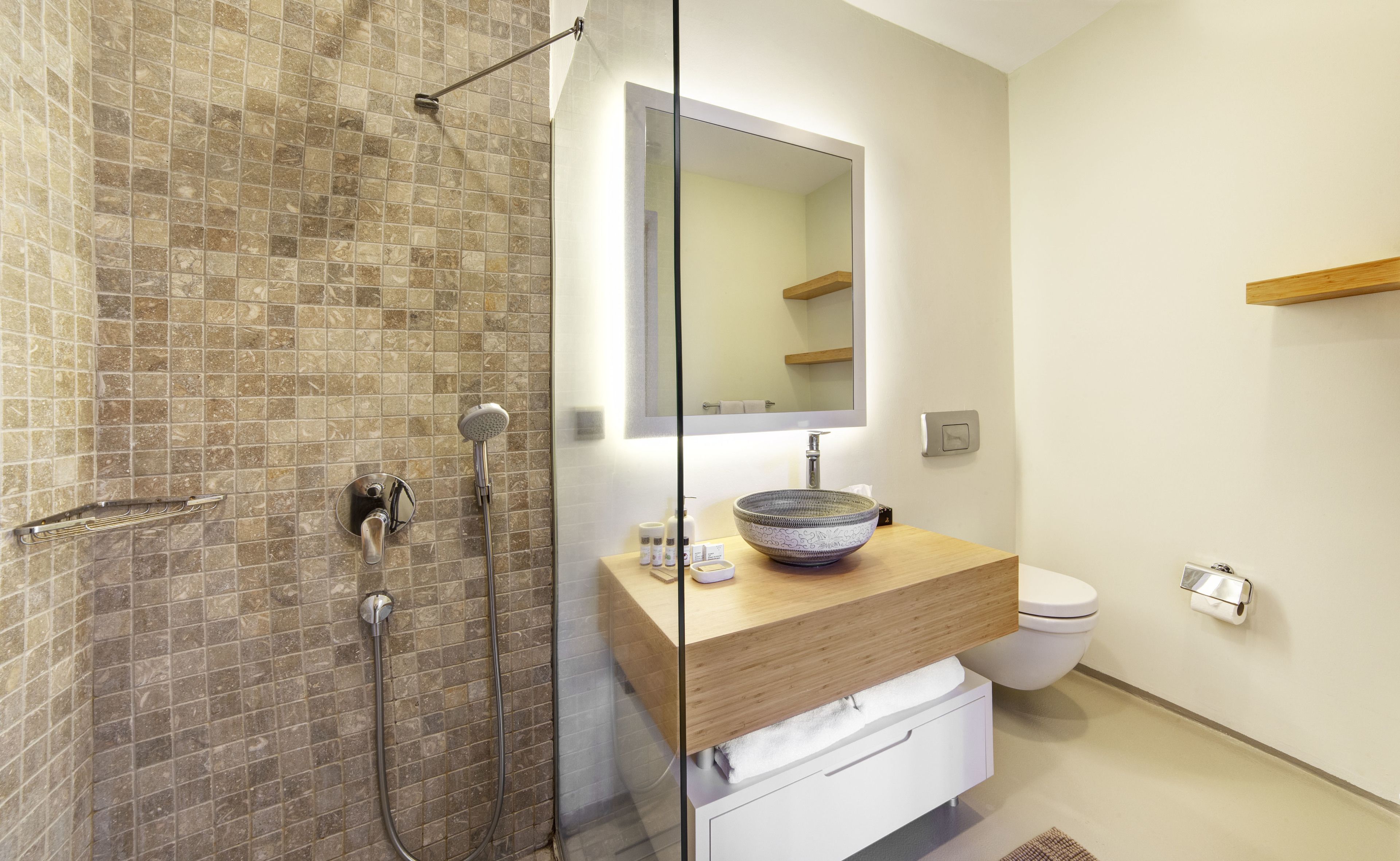 Un baño con cabina de ducha de vidrio, tocador de madera con lavabo a la vista, inodoro y espejo sobre el lavabo.
