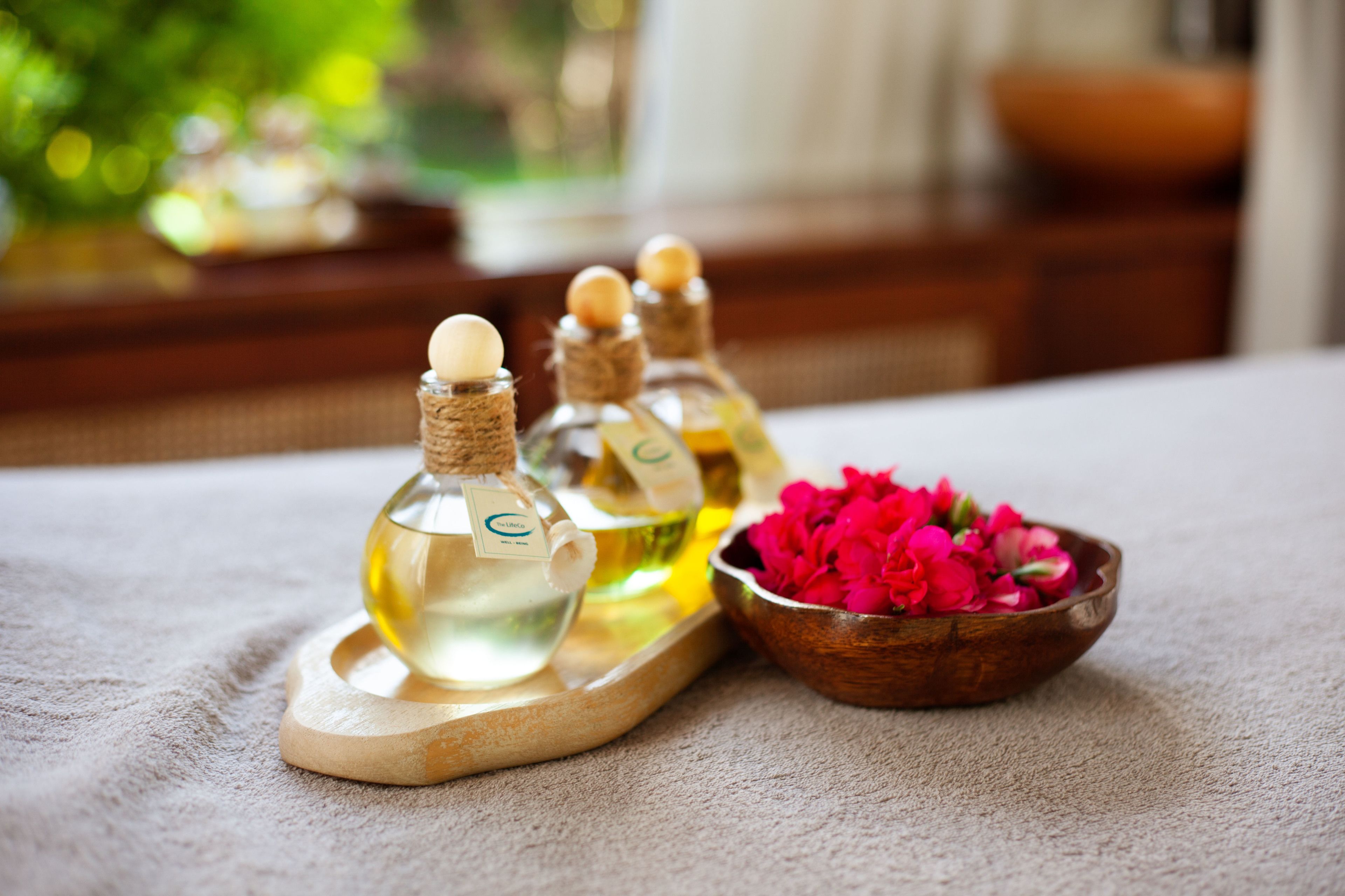 Una bandeja de madera con tres botellas de líquido, un cuenco de madera con flores rosadas, y un fondo borroso con una ventana y follaje.