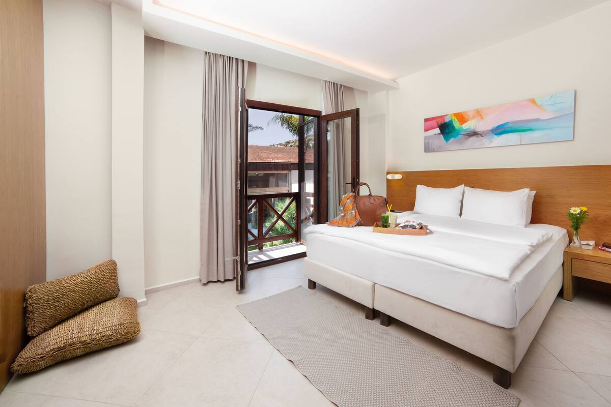 Une chambre d’hôtel avec un lit, une table de chevet, une fenêtre avec des rideaux, un tableau au mur et une vue sur l’extérieur.