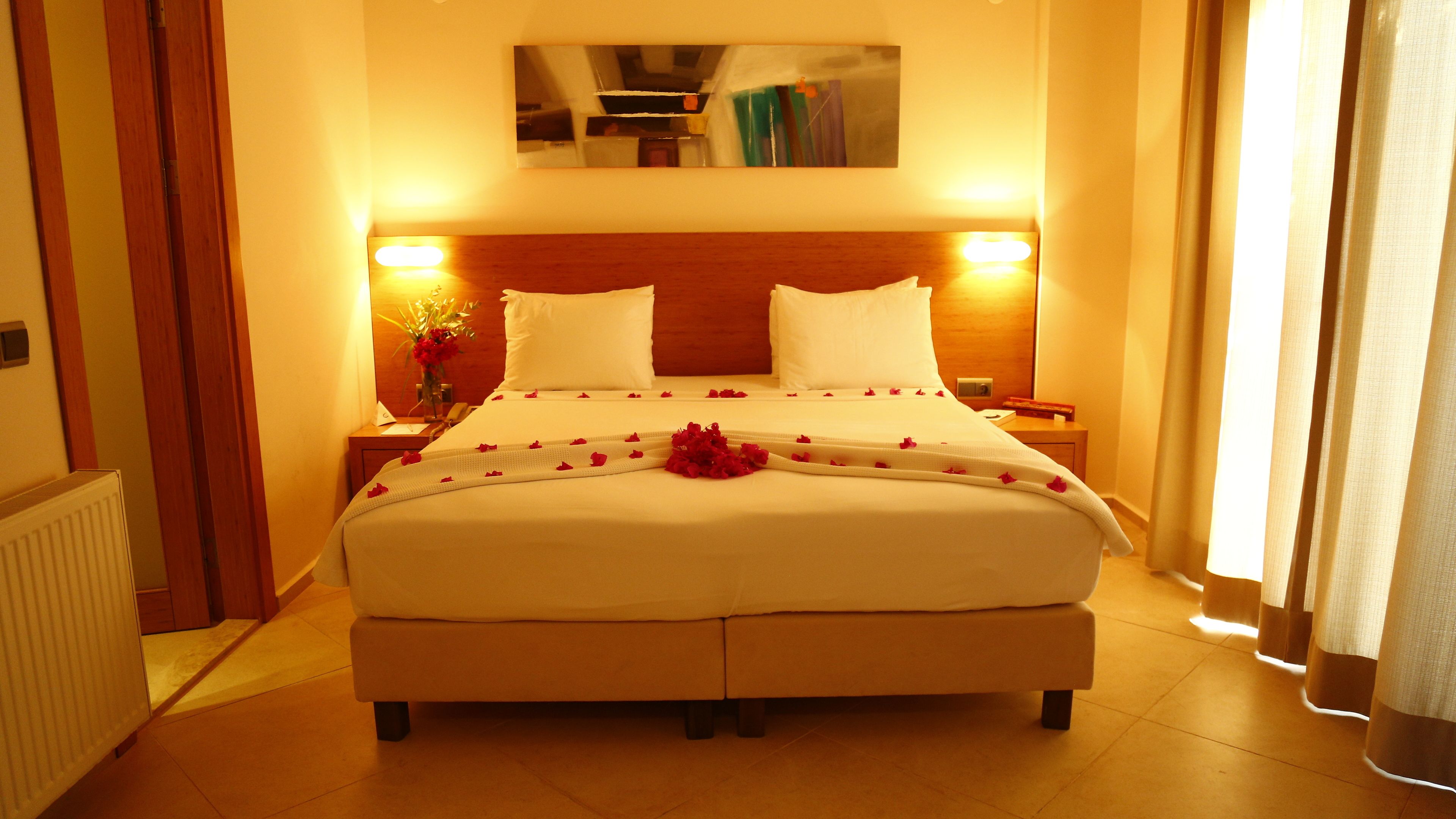 A neatly made bed with white linens and rose petals, a bedside table with a lamp, and a mirror on the wall.
