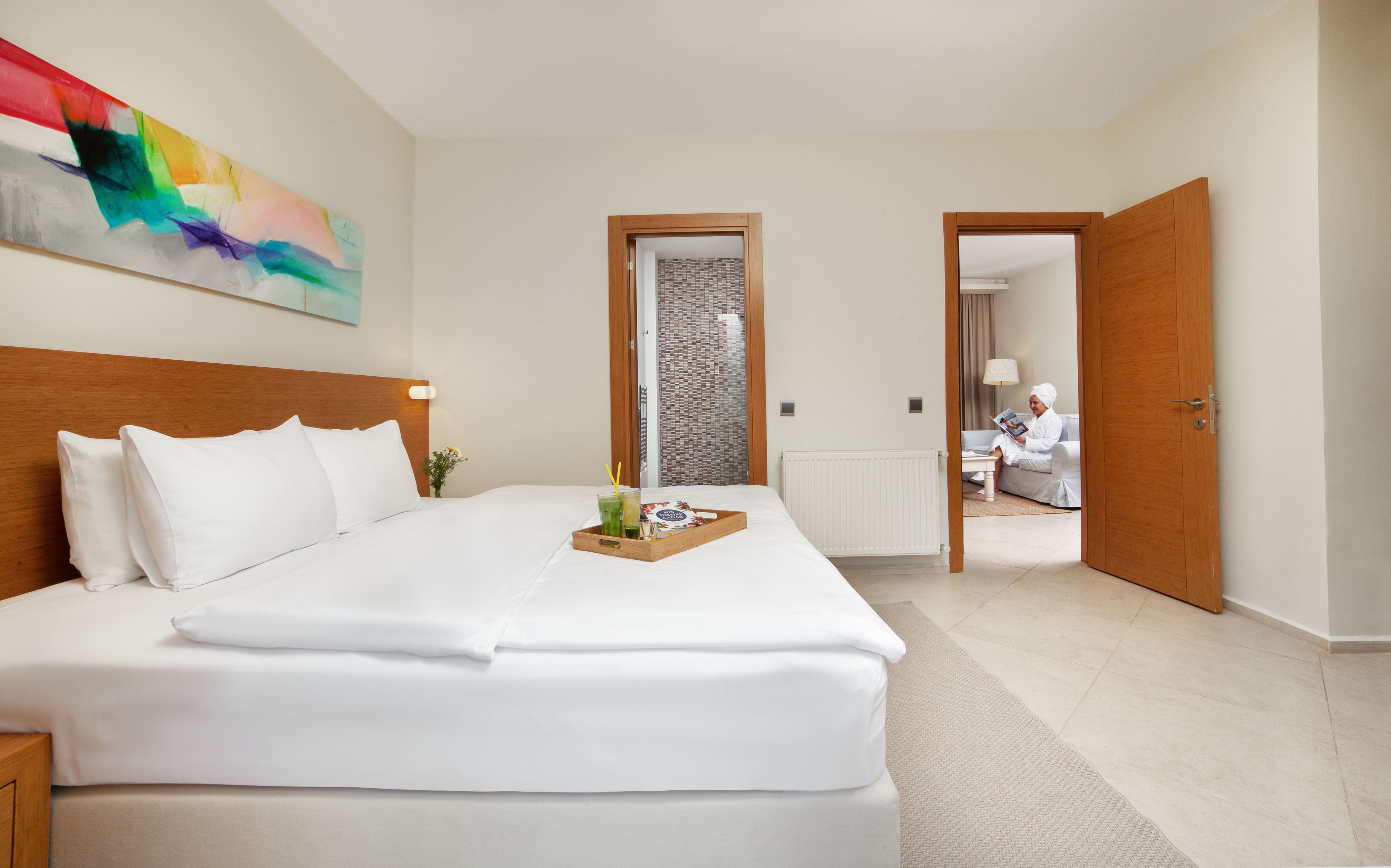 Habitación de hotel con una cama grande, cabecera de madera, mesita de noche con una bandeja de desayuno y un cuadro abstracto colorido en la pared.
