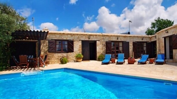 Un espace aménagé au bord de la piscine, avec un bâtiment en pierre, des chaises longues et un ciel d’un bleu limpide.