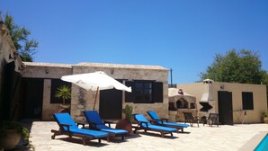 Un espace aménagé au bord de la piscine, avec des bâtiments en pierre, des chaises longues bleues et un parasol.