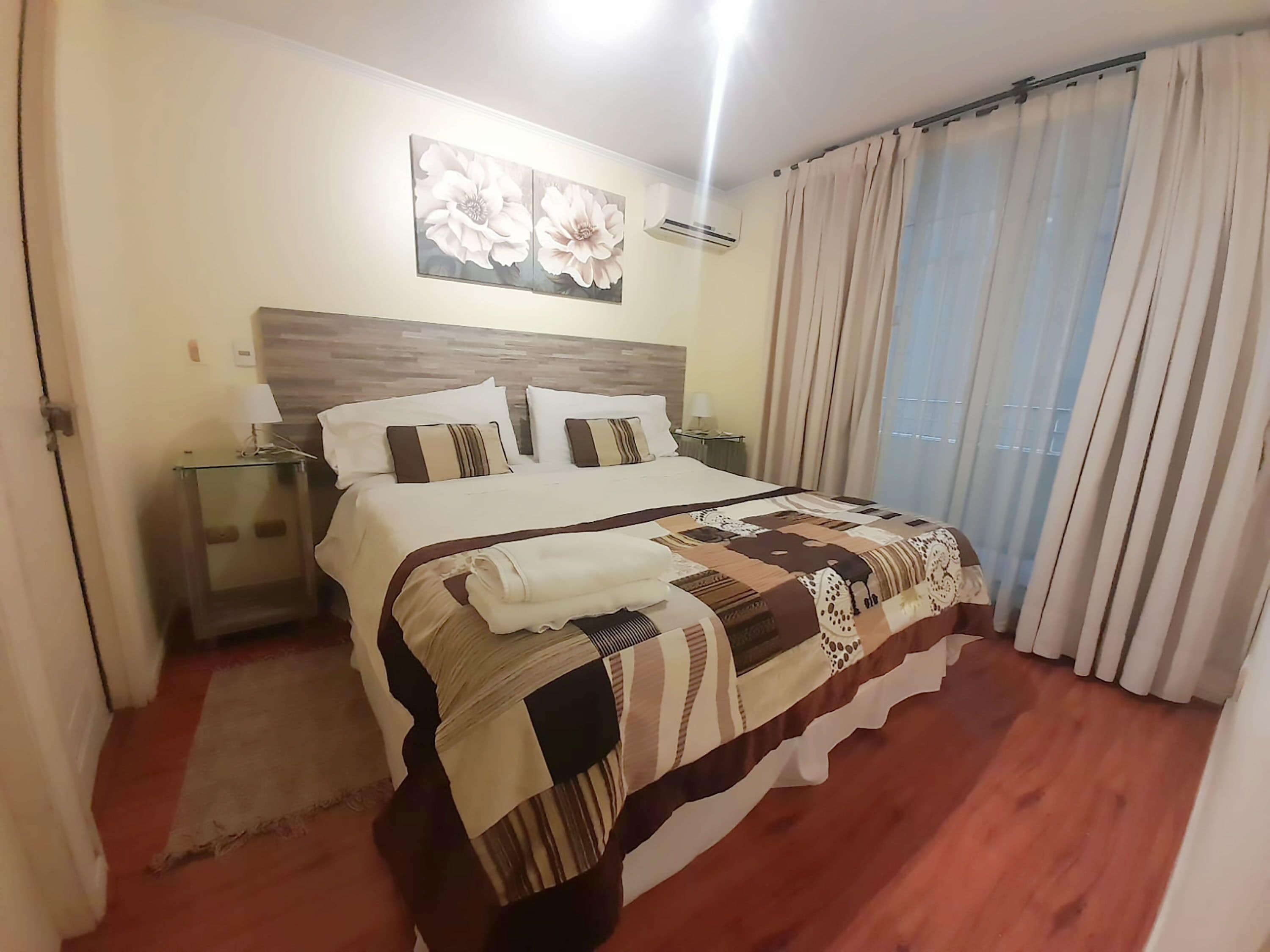 A hotel room with a wooden floor, a large bed with a patterned bedspread, a nightstand with a lamp, a wall-mounted air conditioner, and a window with curtains.